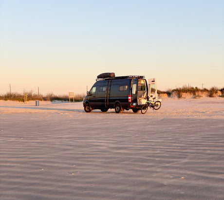 Enjoy an **rv rental** on the beach! Dark camper van with open doors, bike, & roof storage on wavy sand at golden hour.