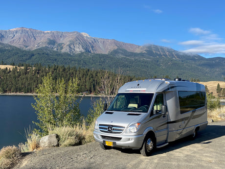 This sleek silver RV rental, a camper van, is parked beside a pristine lake surrounded by lush forests & mountains.