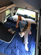 Inside a comfy RV rental, a person reads "Wendell Berry" in a cozy camper bed, next to a small table.