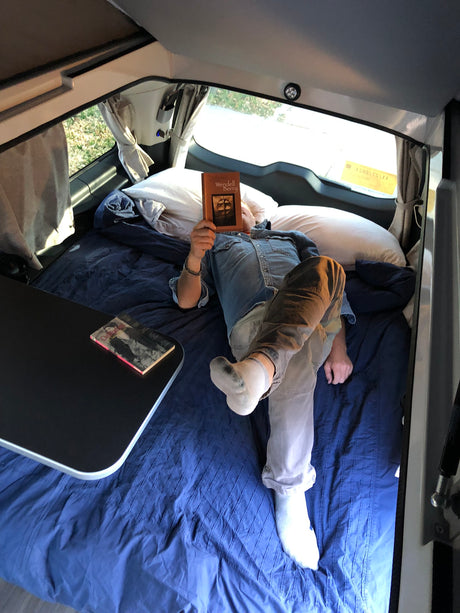 Inside a comfy RV rental, a person reads "Wendell Berry" in a cozy camper bed, next to a small table.
