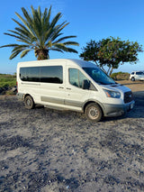 A white van, likely an RV rental, is parked on a gravel lot with a palm tree under a clear blue sky.