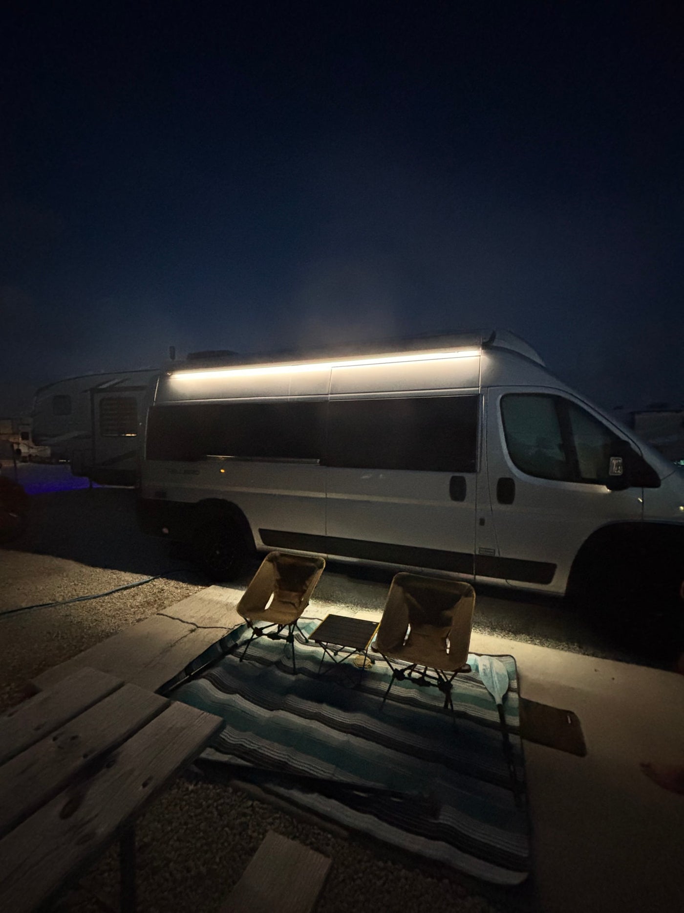Experience an RV rental: a white campervan at night, illuminated by a side light, with two chairs, a small table, and a blanket. — photo 2
