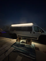 Experience an RV rental: a white campervan at night, illuminated by a side light, with two chairs, a small table, and a blanket.