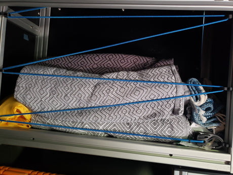 RV rental storage: Patterned blankets neatly secured with blue bungee cords inside a cabinet, preventing items from shifting.