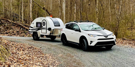 White SUV towing a teardrop RV rental on a gravel road through a bare forest. Ready for outdoor adventure.