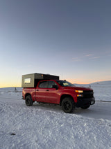 A red Chevy pickup truck with a gray truck camper on a snowy road at sunset. Explore winter landscapes with this RV rental.