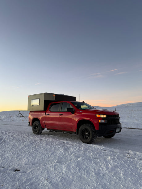 A red Chevy pickup truck with a gray truck camper on a snowy road at sunset. Explore winter landscapes with this RV rental.