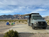 Grey Mercedes-Benz Sprinter camper van (likely rv rental) with extended awning at a desert campsite. Mountains in background, chairs set out.