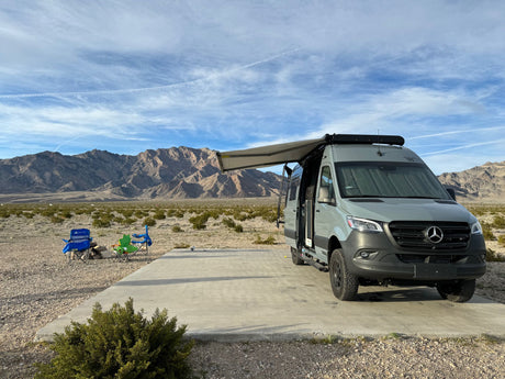 Grey Mercedes-Benz Sprinter camper van (likely rv rental) with extended awning at a desert campsite. Mountains in background, chairs set out.