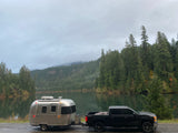 RV rental: A silver Airstream trailer hitched to a truck, parked by a calm lake with forested, misty mountains.