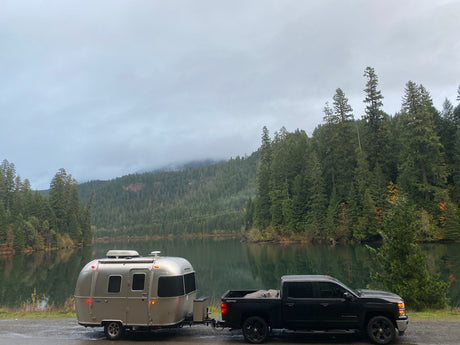 RV rental: A silver Airstream trailer hitched to a truck, parked by a calm lake with forested, misty mountains.