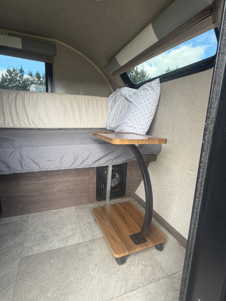 RV rental interior: A cozy bed with a pillow, a window showing trees, and a wooden rolling table on wheels.