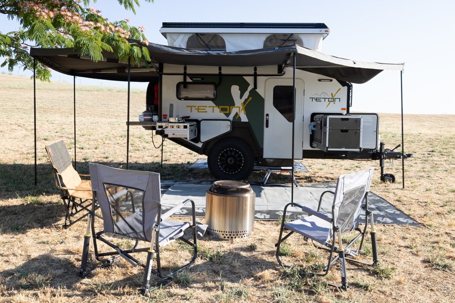 An off-grid TetonX rv rental trailer, fully deployed with a rooftop tent, awning, outdoor kitchen, chairs, and fire pit in a field. — photo 12
