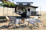 An off-grid TetonX rv rental trailer, fully deployed with a rooftop tent, awning, outdoor kitchen, chairs, and fire pit in a field.