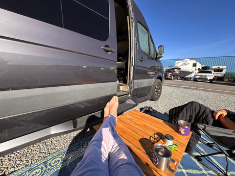 Feet up, relaxing outside a gray rv rental camper van at a sunny campsite with drinks on a table.