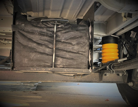 Underside of an RV rental showing a mounted battery box and a bright yellow suspension helper spring.
