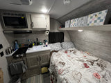 Cozy RV rental interior: compact kitchen with microwave, stove, sink, and a bed featuring a unique postcard-themed comforter.