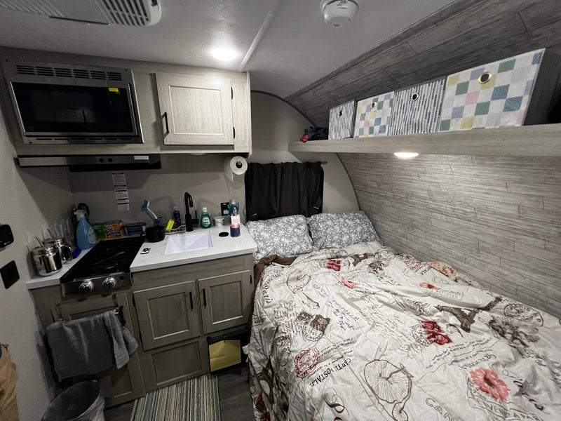Cozy RV rental interior: compact kitchen with microwave, stove, sink, and a bed featuring a unique postcard-themed comforter. — photo 8