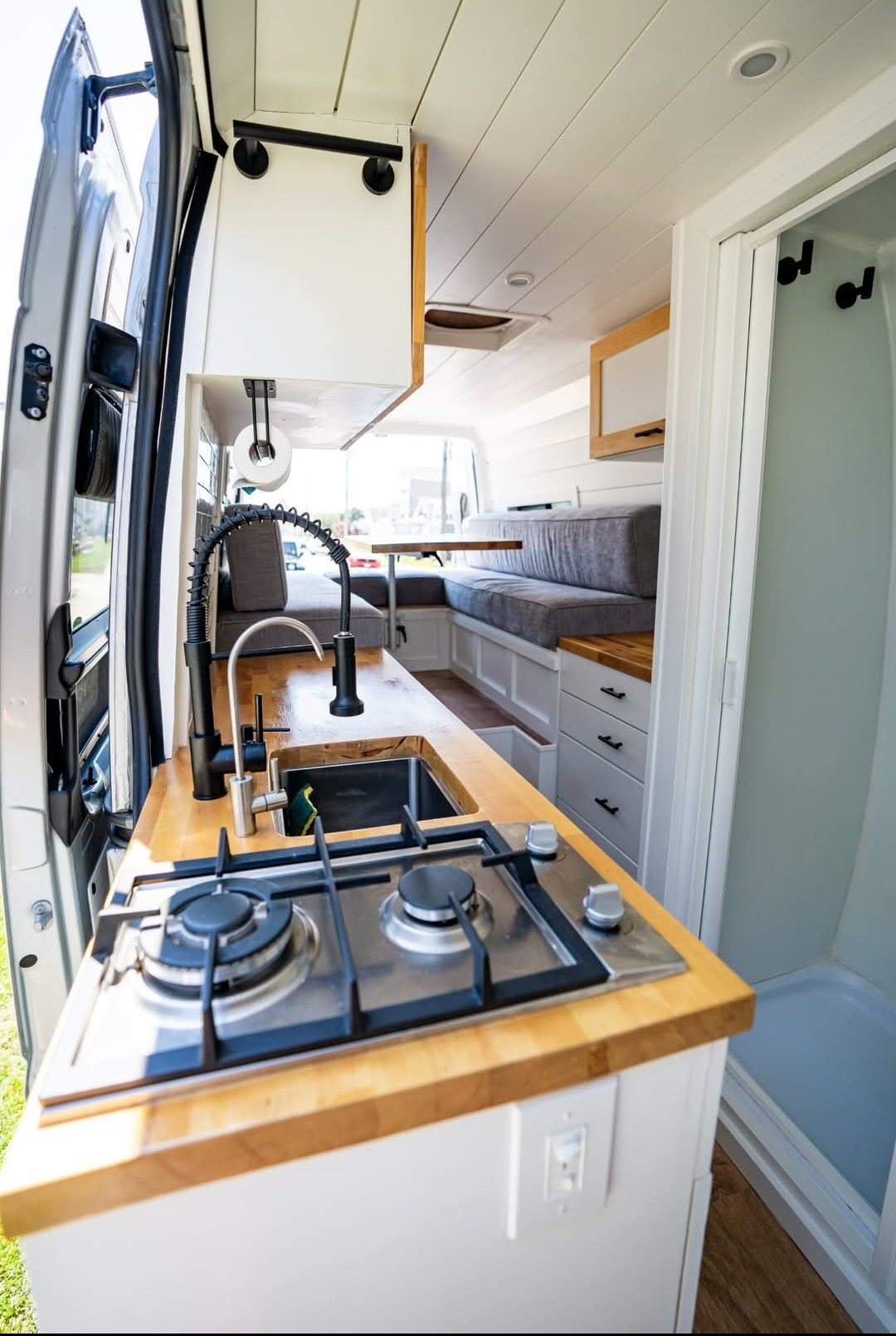 Bright rv rental campervan interior with a compact kitchen: stove, sink, and wood counters. A dinette and shower stall are also visible. — photo 4