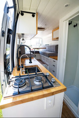Bright rv rental campervan interior with a compact kitchen: stove, sink, and wood counters. A dinette and shower stall are also visible.