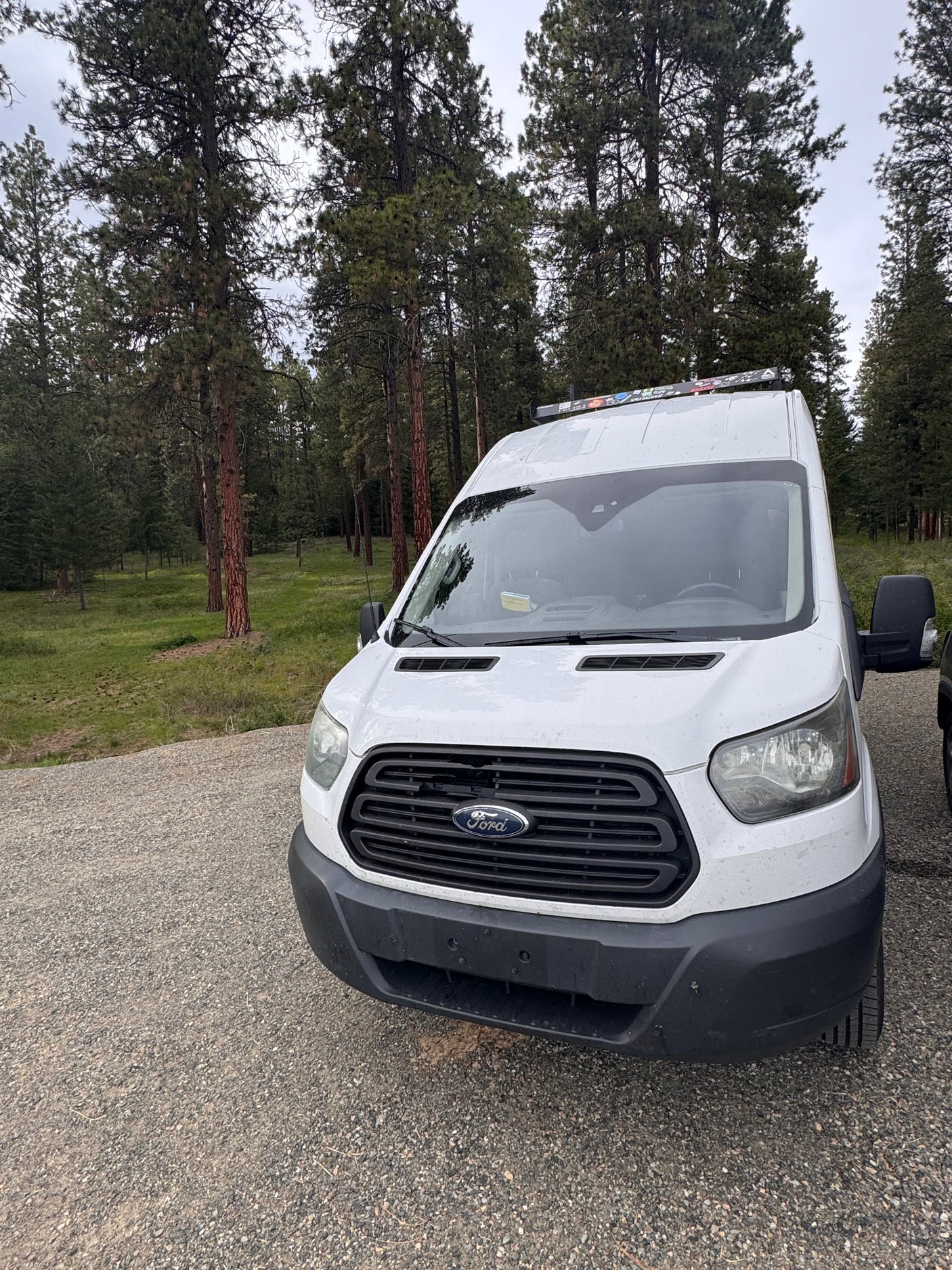 This white Ford Transit camper van, ideal for an RV rental, is parked on a gravel road by a peaceful forest.
