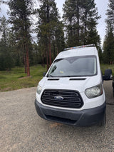 This white Ford Transit camper van, ideal for an RV rental, is parked on a gravel road by a peaceful forest.