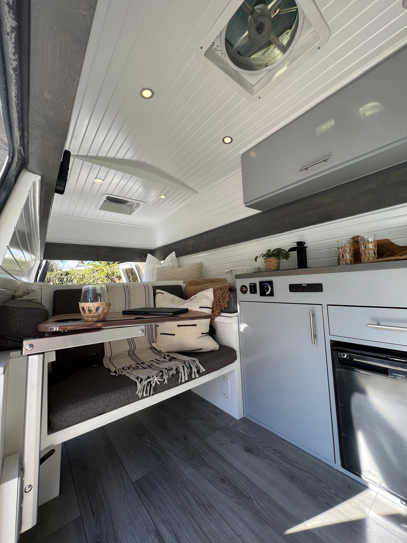Modern RV rental interior: white slatted ceiling, gray kitchenette with fridge, cozy bench seating & table. Adventure awaits!
