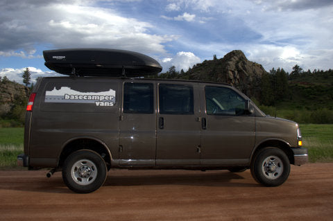 Brown Basecamper van, ideal for RV rental. Equipped with roof box, on a scenic dirt road with mountains.