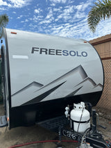 FREESOLO teardrop RV rental, grey with mountain graphics, propane tank, under a cloudy blue sky.