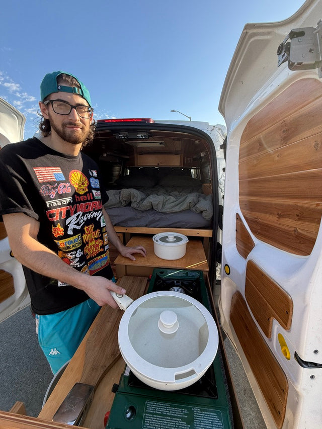 Cooking outside a custom RV rental van, showcasing its cozy bed and outdoor kitchen setup.
