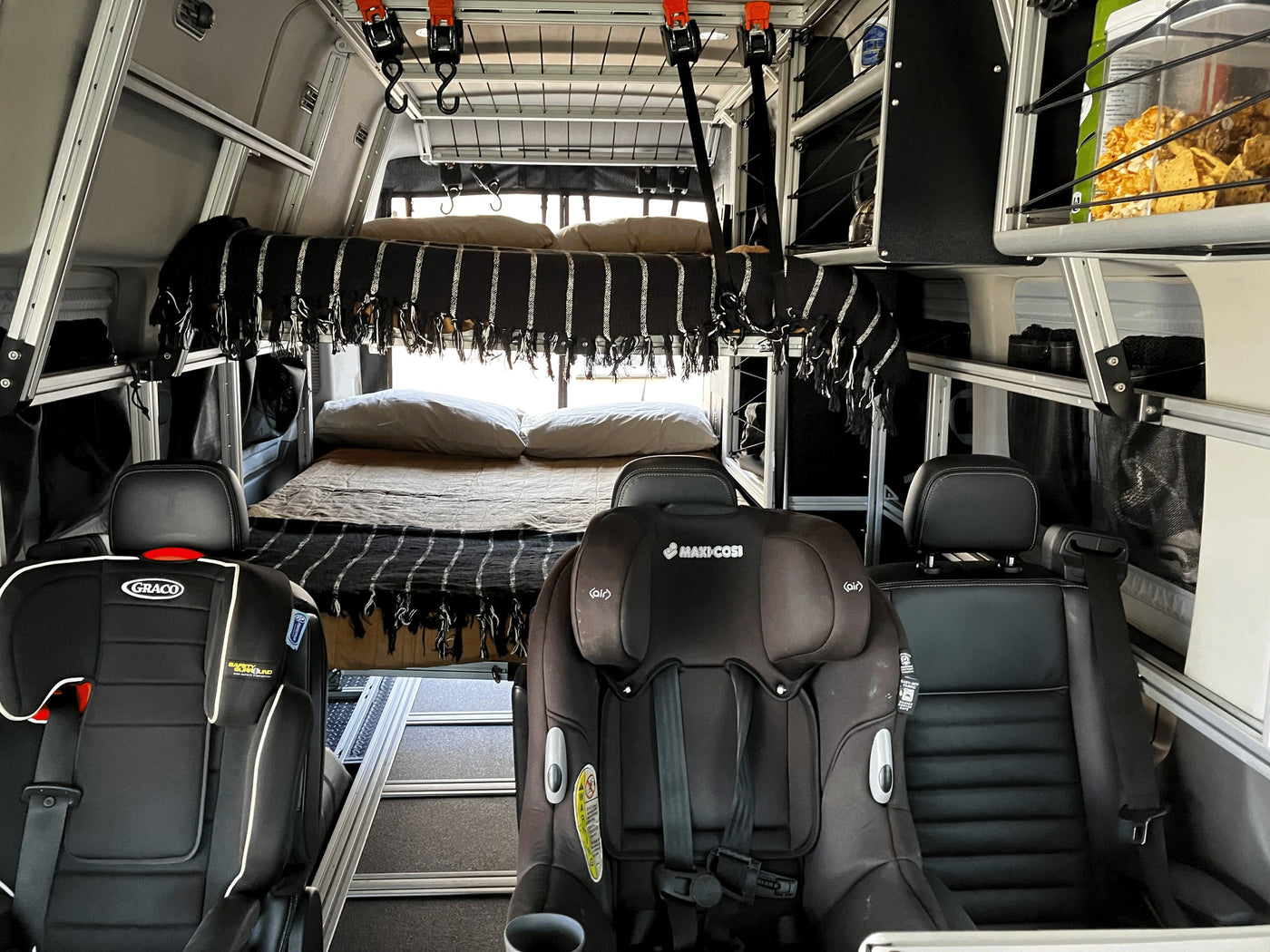 Family-friendly RV rental: A camper van interior features a cozy bed, two child car seats (Graco & Maxi-Cosi), and smart storage. — photo 20
