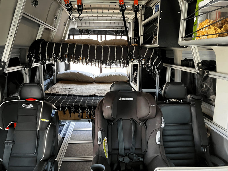 Family-friendly RV rental: A camper van interior features a cozy bed, two child car seats (Graco & Maxi-Cosi), and smart storage. — photo 20