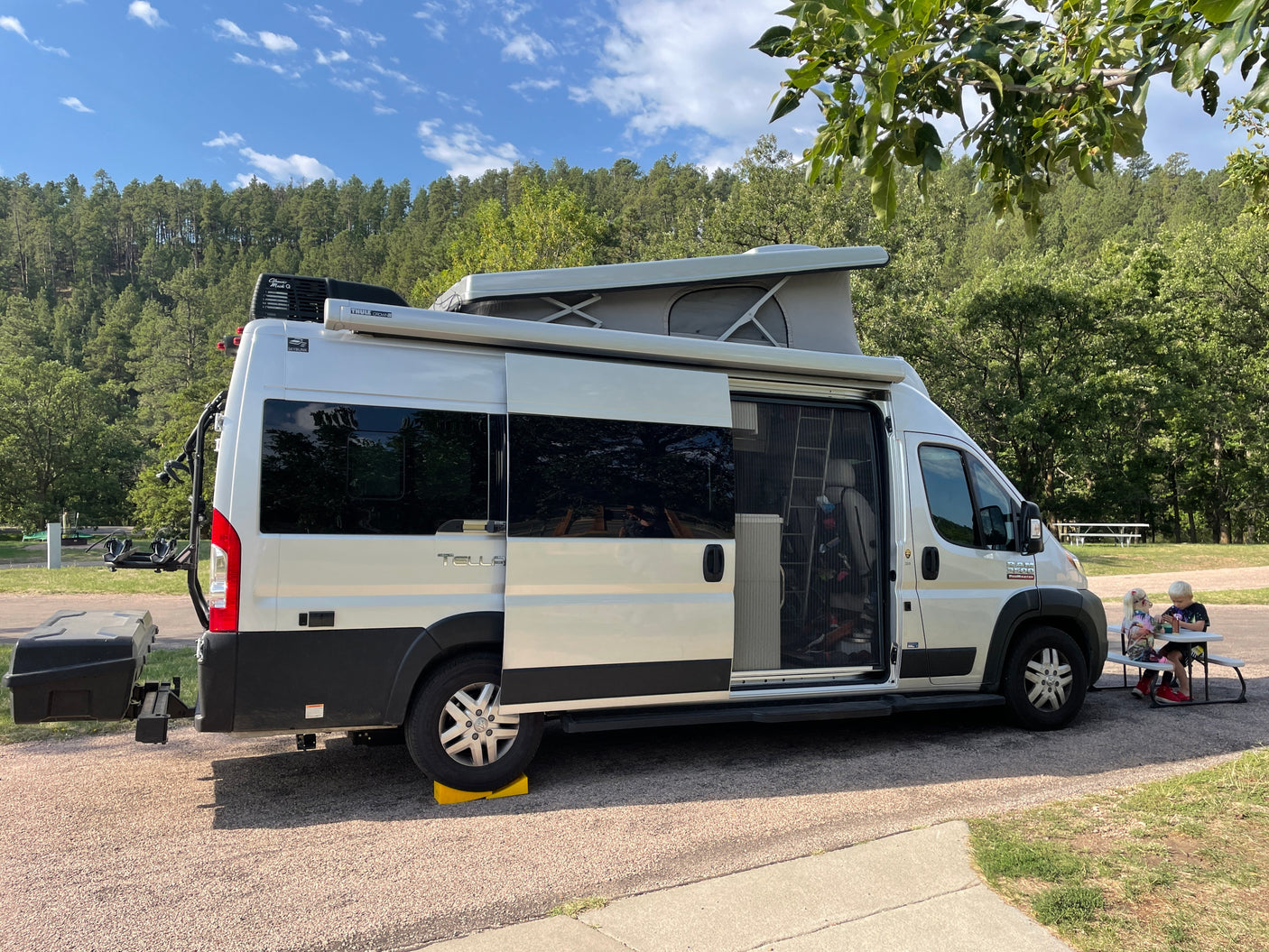 A silver camper van with a pop-top, an ideal rv rental, is parked at a wooded campsite as kids eat at a picnic table.