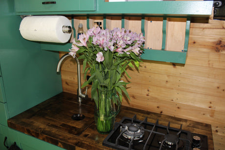 Inside an RV rental: a compact kitchen with a sink, stove, paper towels, and pink alstroemeria flowers.