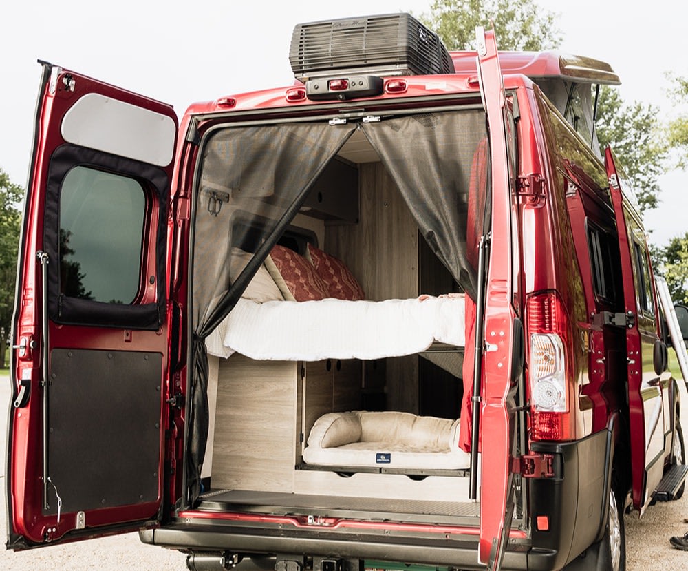 Red camper van RV rental interior: upper bed, lower pull-out pet bed, and bug screen at open rear doors.