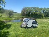 Enjoy nature with an RV rental! A grey teardrop camper is parked by a sunny stream in a green meadow, surrounded by trees.