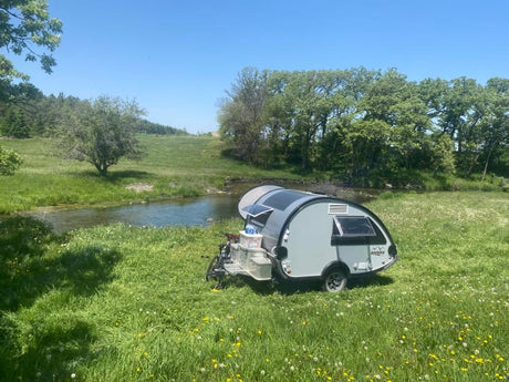 Enjoy nature with an RV rental! A grey teardrop camper is parked by a sunny stream in a green meadow, surrounded by trees.