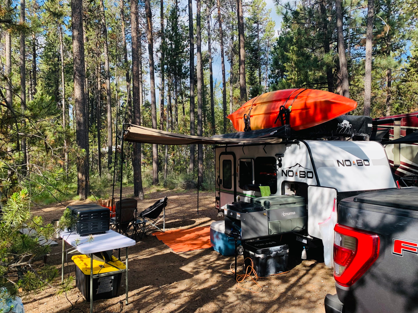 Forest campsite with a white adventure trailer, orange kayak, and outdoor setup. Ideal for an RV rental getaway.