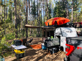 Forest campsite with a white adventure trailer, orange kayak, and outdoor setup. Ideal for an RV rental getaway.