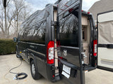 Dark camper van (RV rental) parked on concrete, rear doors open revealing interior, with electric hookup.