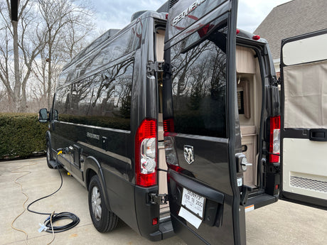 Dark camper van (RV rental) parked on concrete, rear doors open revealing interior, with electric hookup.