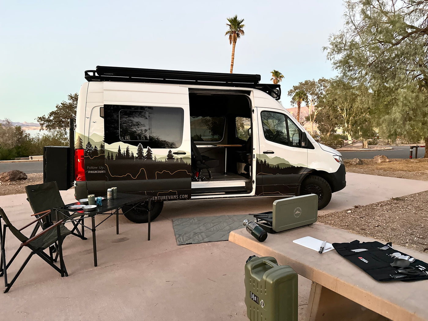 RV rental van setup for outdoor adventure: a white camper van with camping gear, table, chairs, and stove by a lake. — photo 12