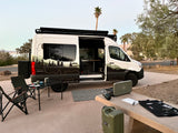 RV rental van setup for outdoor adventure: a white camper van with camping gear, table, chairs, and stove by a lake.