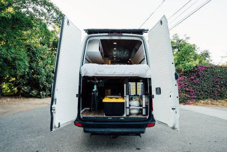 Rear view of an RV rental camper van with open doors, showcasing a bed, bike, and storage space for gear.