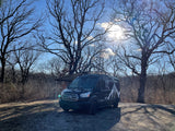 This black camper van, likely an RV rental, is parked with its awning extended amidst bare trees on a sunny day.