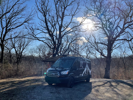 This black camper van, likely an RV rental, is parked with its awning extended amidst bare trees on a sunny day.