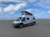 Silver camper van RV rental parked on a vast sandy beach with the ocean in the background under a cloudy sky.