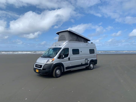 Silver camper van RV rental parked on a vast sandy beach with the ocean in the background under a cloudy sky.