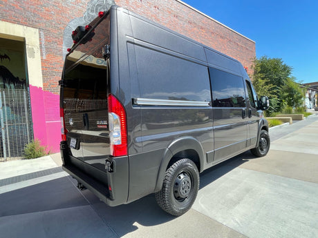 Dark grey Ram ProMaster van (an RV rental) with open rear doors, revealing its conversion, parked by a brick wall.