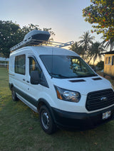 This white Ford Transit rv rental, featuring a roof rack, is parked on green grass at sunset, ideal for island camping.